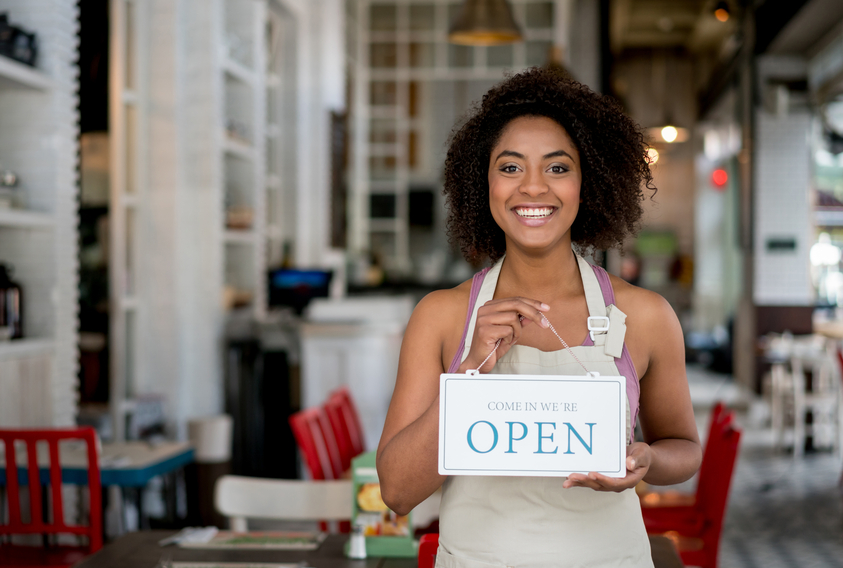 Happy African American waitress holding an open sign at a restaurant and looking at the camera smiling