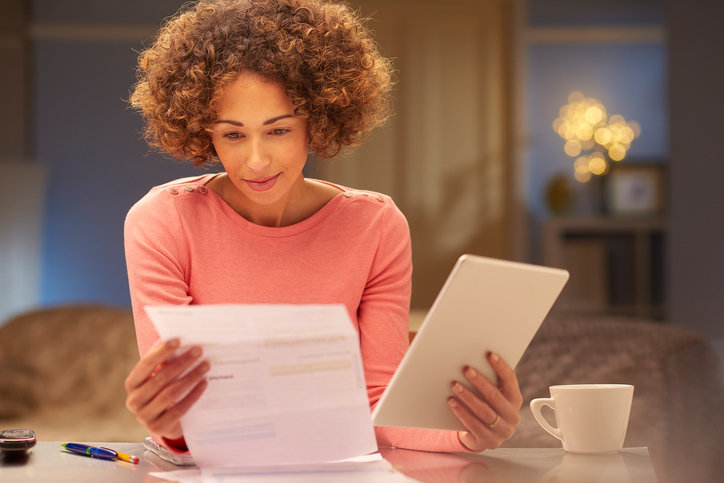 a young woman sits at home at night , going through her domestic bills and comparing them with comparison websites using her digital tablet .