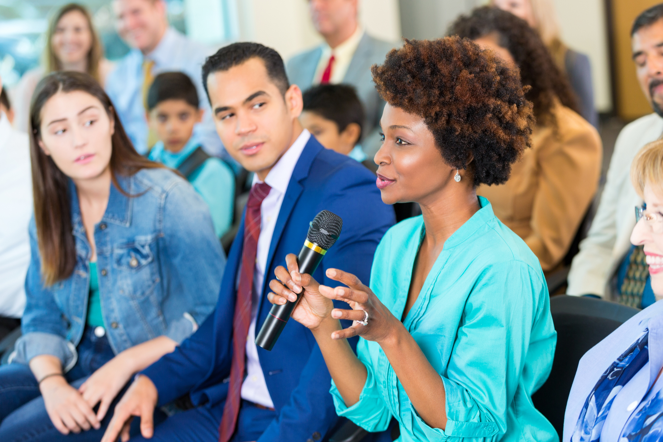 Beautiful mature African American woman gestures while asking a political candidate a question during a town hall meeting. A diverse group of people are sitting with her in the audience.