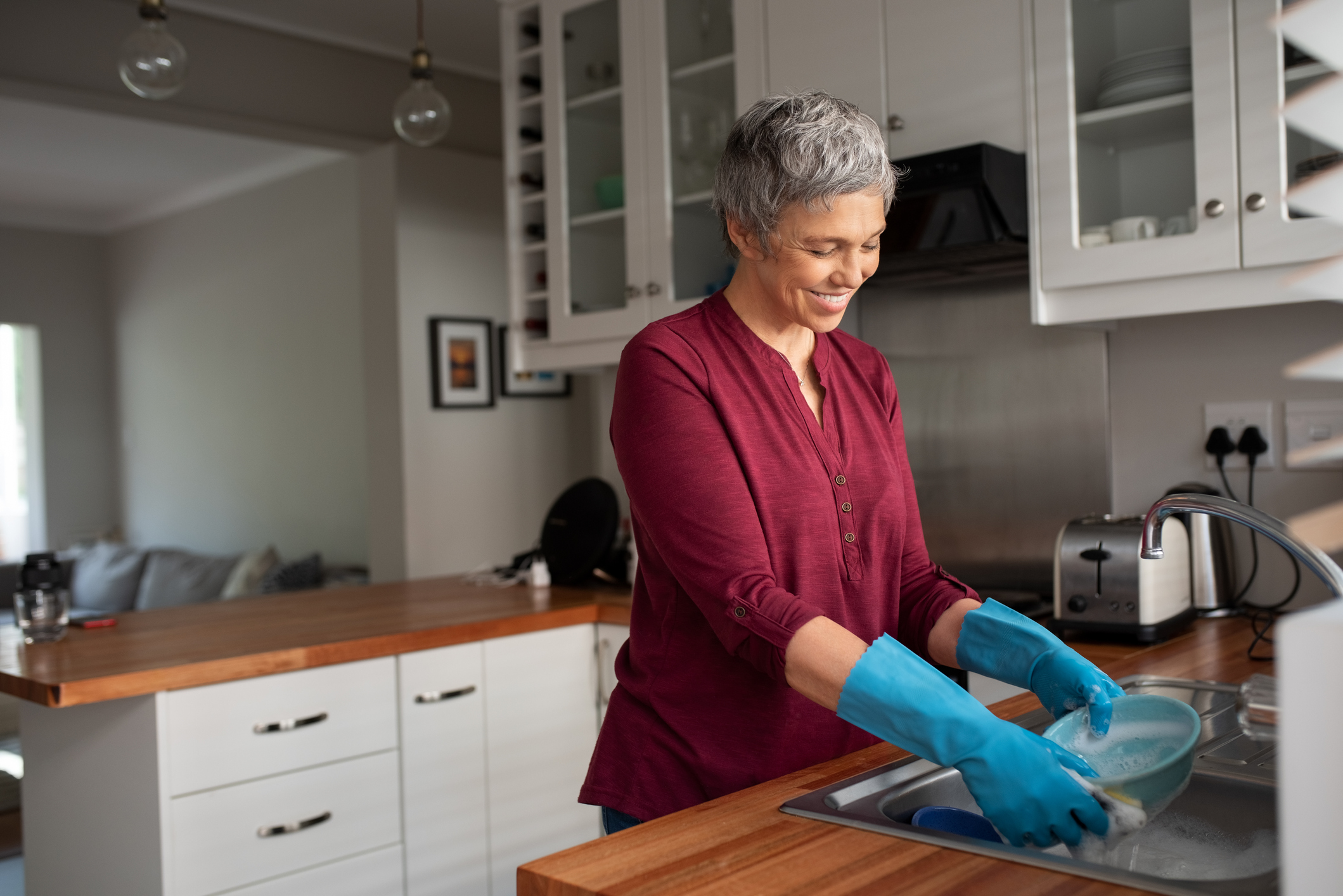 Senior woman washing dishes in her kitchen. Cheerful mature housewife in kitchen. Beautiful smiling woman with gray hair and blue gloves washing bowl in washbasin at home.