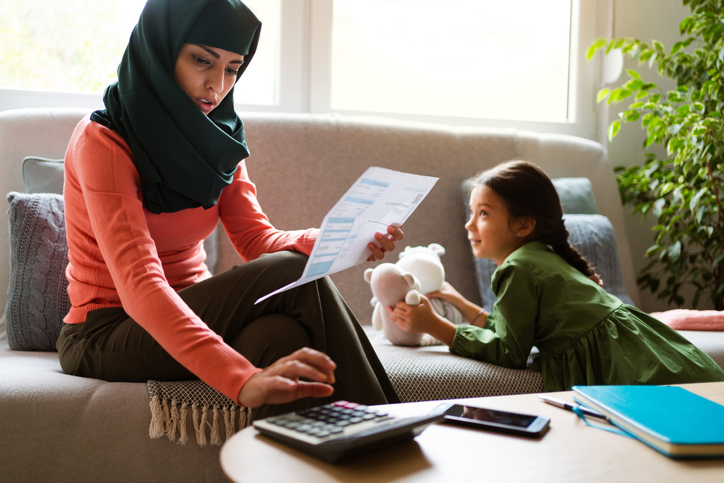 Young Muslim woman calculating home finances, paying bills while her daughter is playing with toys