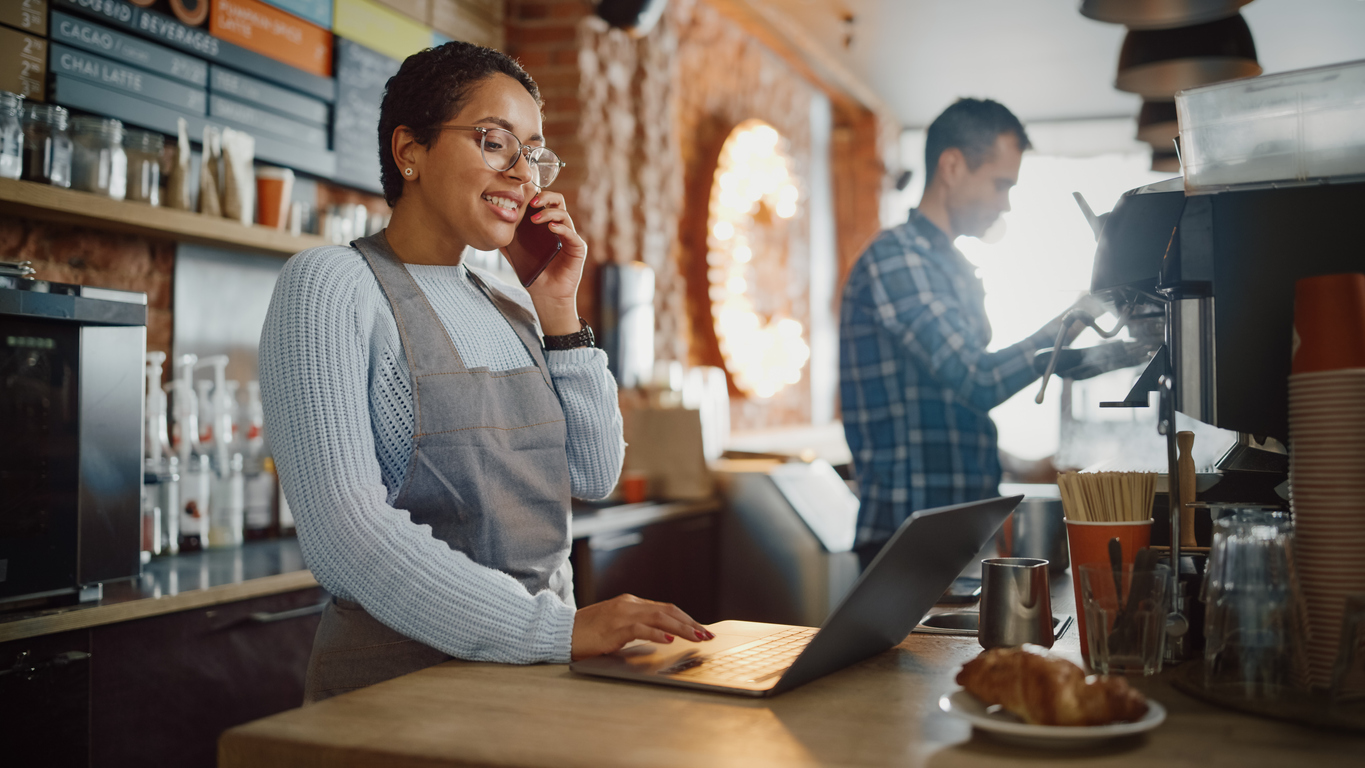 Latin American Coffee Shop Employee Accepts a Pre-Order on a Mobile Phone Call and Writes it Down on Laptop Computer in a Cozy Cafe. Restaurant Manager Browsing Internet and Talking on Smartphone.