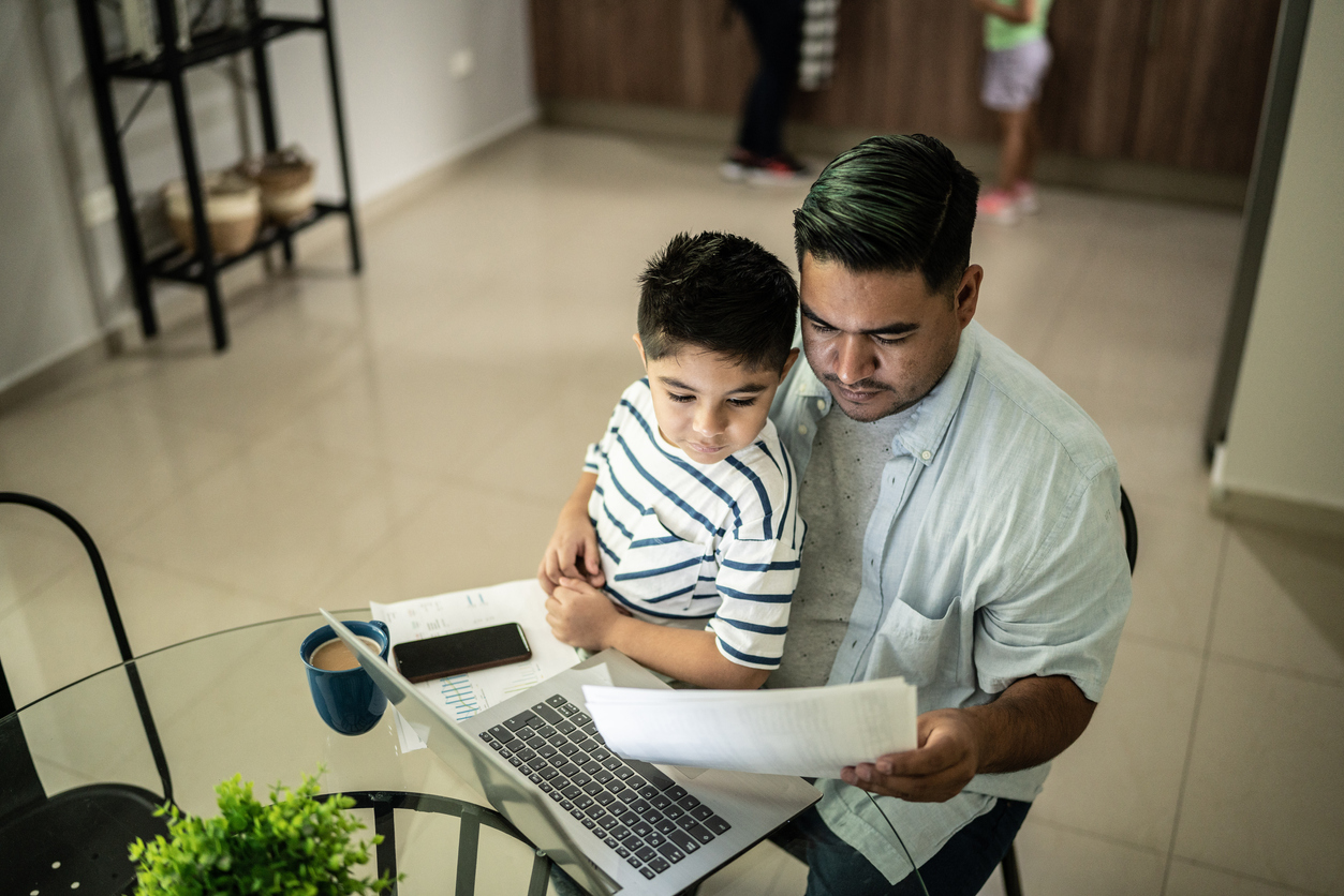 A dad sits with his young son at the laptop looking at a bundle of bills