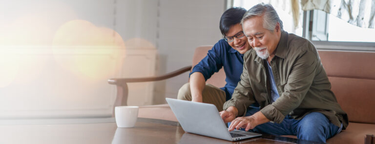 happy asian adult son and senior father sitting on sofa using laptop together at home