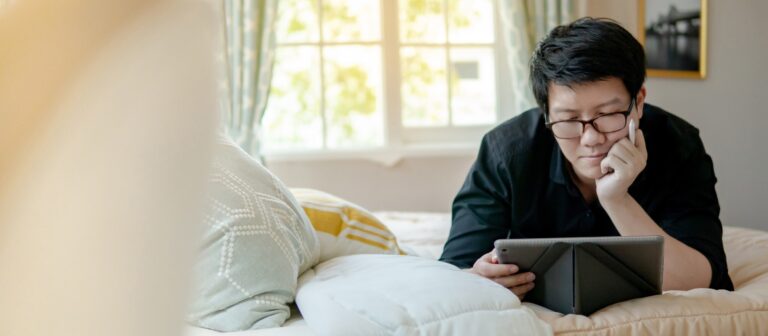 Asian man lying on cozy bed using digital tablet and pen in bedroom.