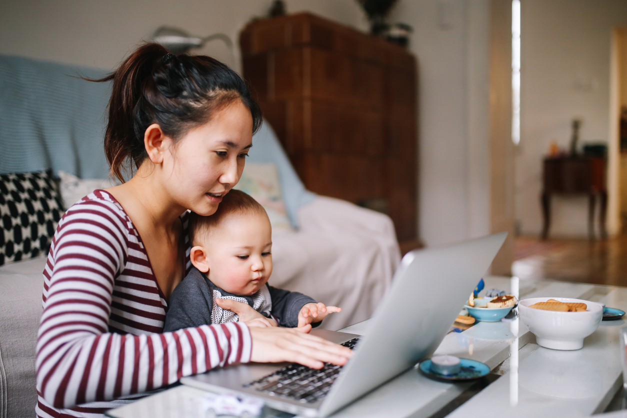 young Asian woman sitting at home, doing some freelance job while taking care of her little baby boy.