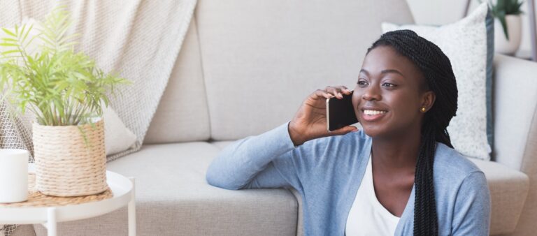 Happy african american woman talking on cellphone with friend while sitting on floor at home