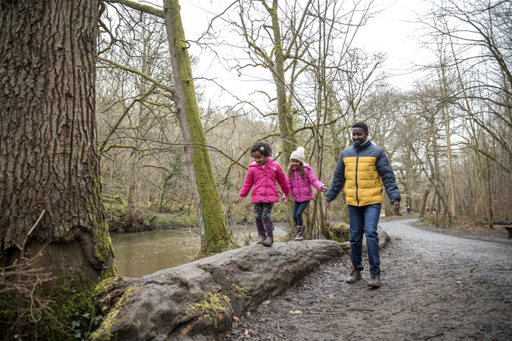 Two girls and man walking in wooded area alongside river water