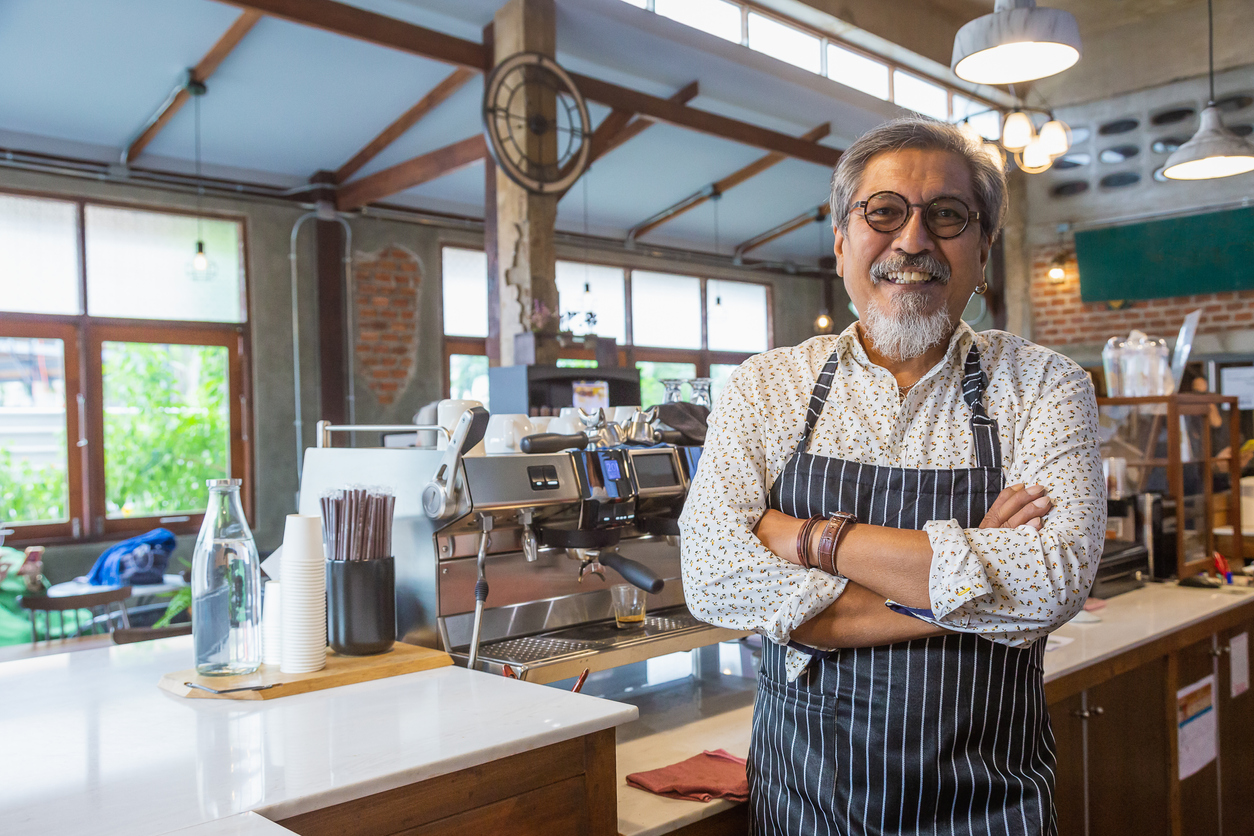 A smiling Asian man stands with his arms folded in his coffee shop
