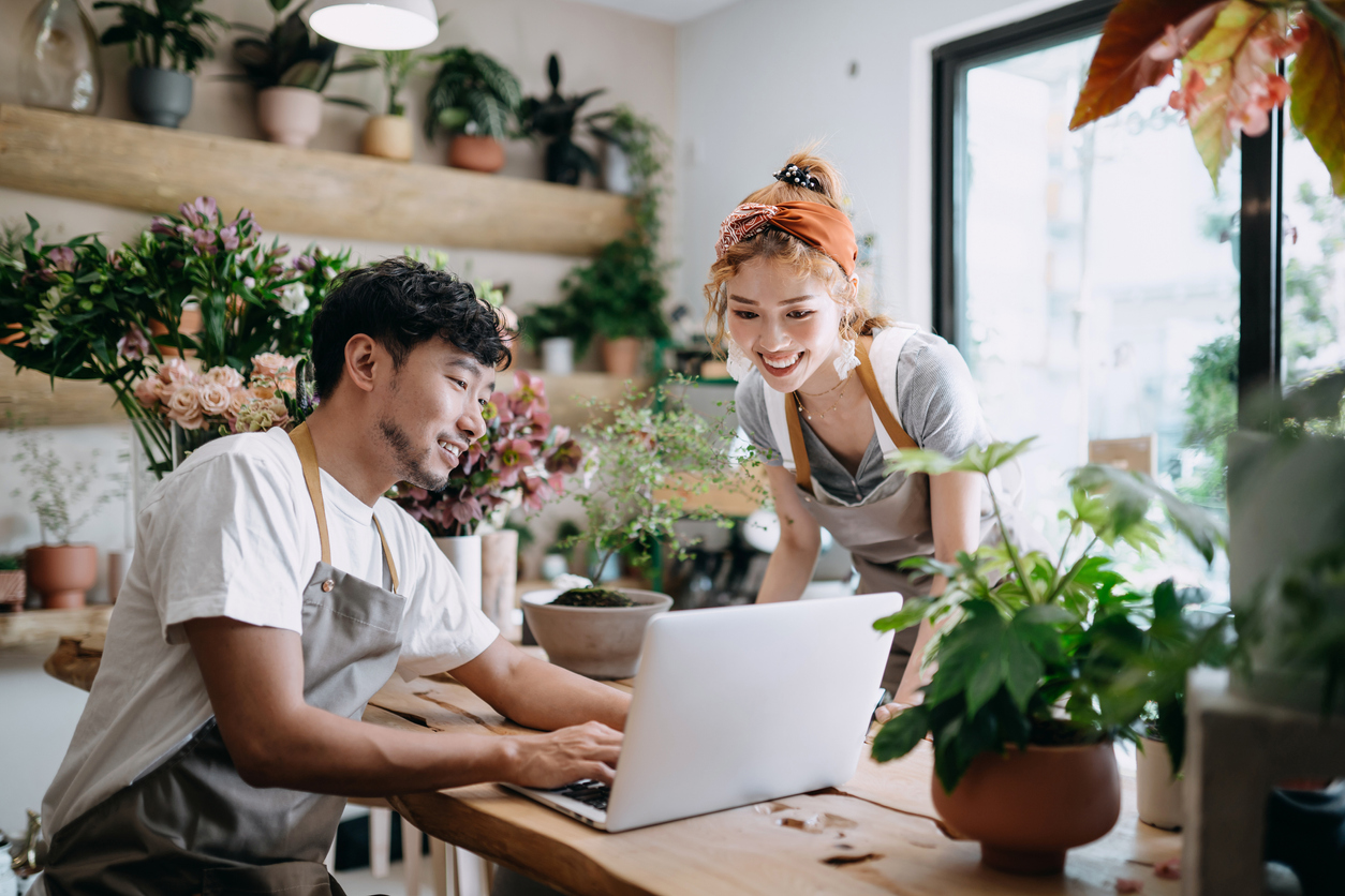 A smiling young Asian couple, the owners of a florist, have a discussion while looking at their laptop.