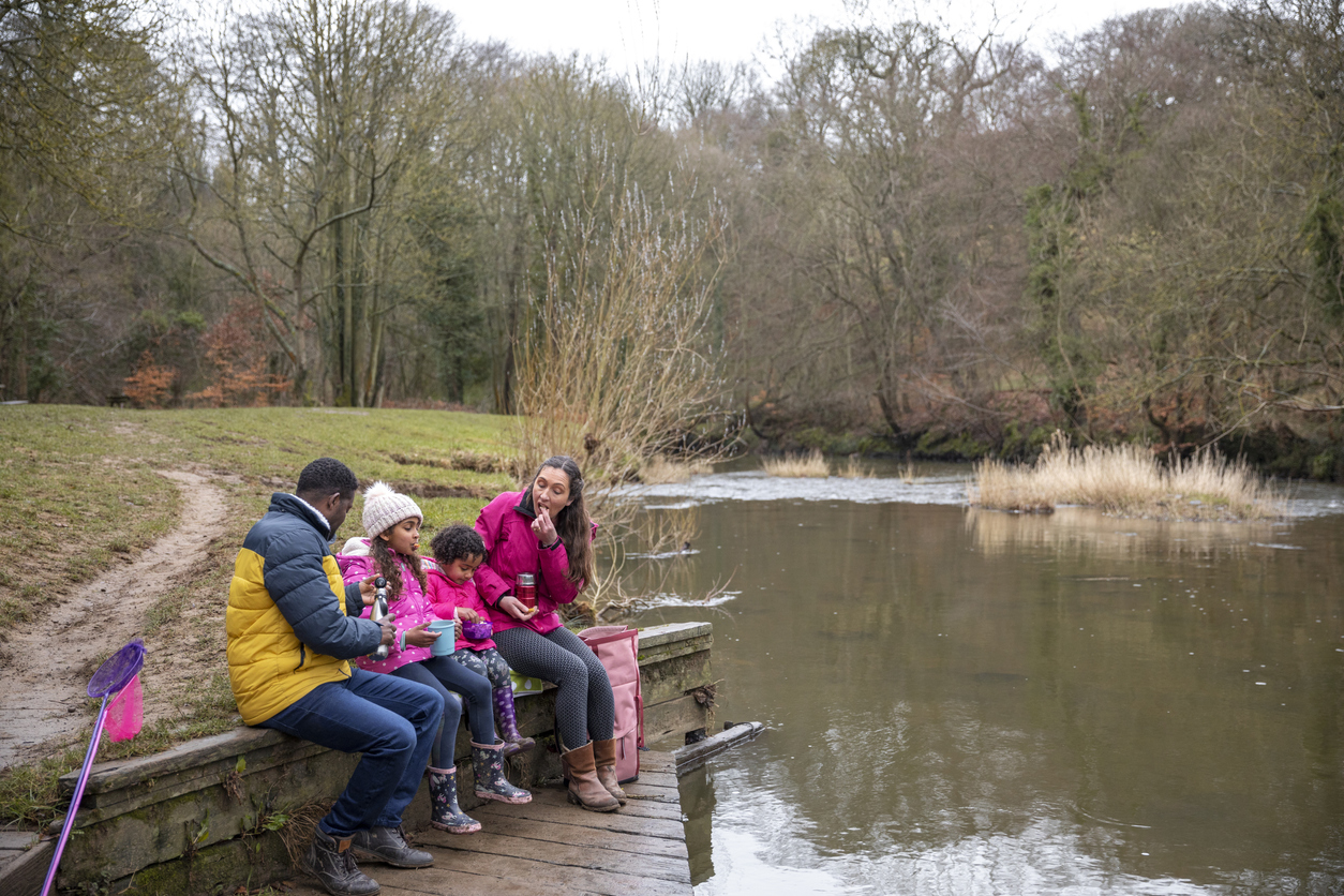 A family are sitting on a wall next to a river. They are taking a break from fishing by having a picnic.