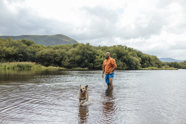 Man standing in a like with his dog running through the water