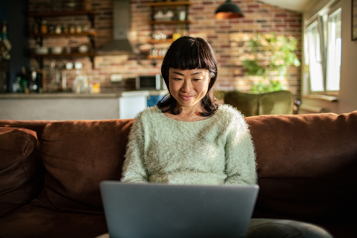 A smiling, young Japanese woman is sat on her sofa using the laptop.