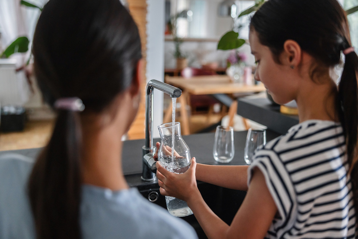 Two young multiracial sisters are standing in front the sink while one is filling a glass jar with some tap water. The other one is standing next to her and observing how she does it.