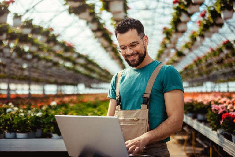 A young male business owner using laptop in greenhouse filled with colorful flowers