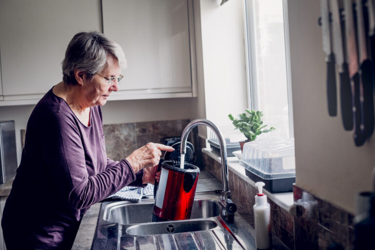 An older woman is filling up a kettle while standing at her kitchen sink.