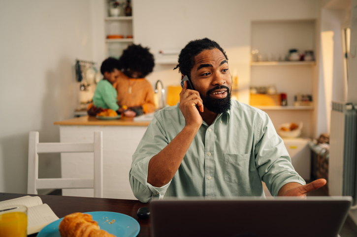Man sitting at the table with a laptop and breakfast on it and having a phone call while his family standing in a kitchen.