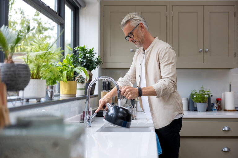 A white middled-aged man wearing glasses is filling up his kettle at the kitchen sink.