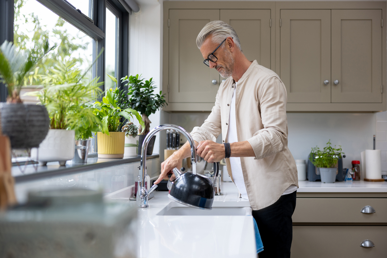A white middled-aged man wearing glasses is filling up his kettle at the kitchen sink.