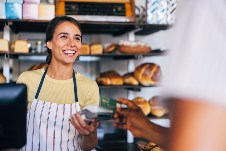 A cheerful baker serves a customer in a warm, artisan bakery setting. Freshly baked bread is displayed in the background, highlighting a friendly and welcoming atmosphere.