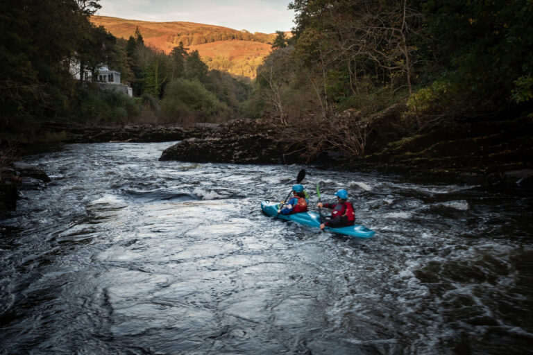 The evening sun is setting on the hills as two people kayak on the River Dee in Wales.