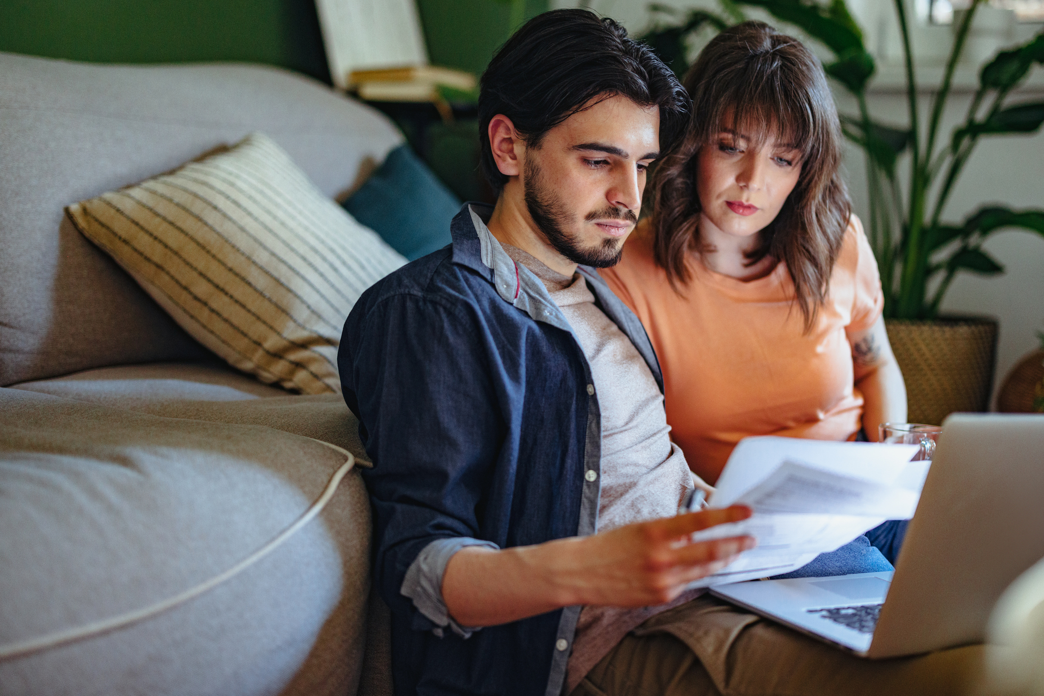 A young couple sit on the floor in front of their sofa looking at bills in front of a laptop.