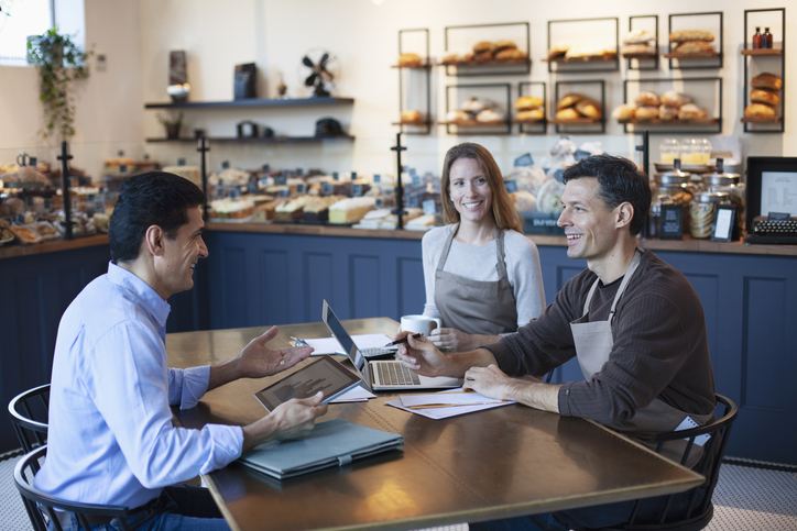 mature man talking advice with mature women and mature man owner staff employee sitting together in small business bakery cafe using laptop computer digital tablet planning