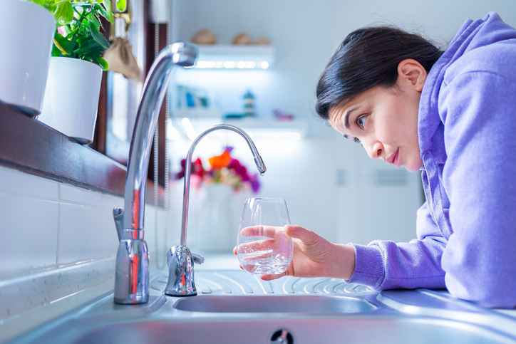 Young and worried woman is waiting for a last drop of water hangs from a tap with glass under water shortage