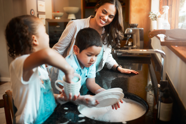 Young happy mixed race mother washing the dishes with her son and daughter in the kitchen at home. Little brother and sister helping their mom with the dishes.